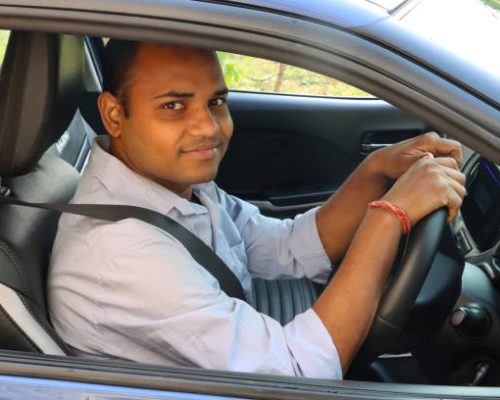 Stock photo showing an Indian man driving in his car (a private hire taxi cab car), wearing a seatbelt and looking out of open driver's window.