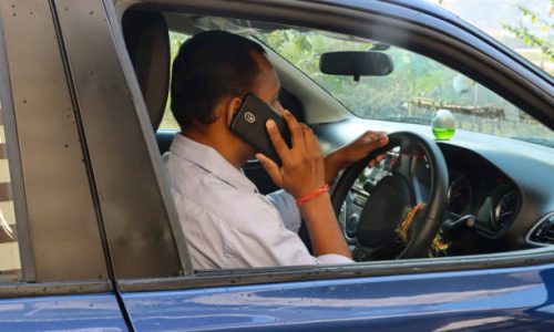 Stock photo showing an Indian man driving in his car (a private hire taxi cab car), whilst talking to a friend on his mobile phone. The driver is pictured without a seatbelt, with a smart phone being held to his ear as his listens and drives. Concept image for dangerous driving, since using a phone behind the wheel is illegal and considered to be both dangerous and a serious traffic offence, punishable with large fines and points on the relevant driving licence.