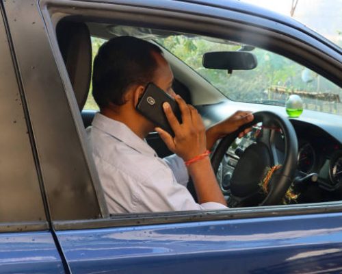 Stock photo showing an Indian man driving in his car (a private hire taxi cab car), whilst talking to a friend on his mobile phone. The driver is pictured without a seatbelt, with a smart phone being held to his ear as his listens and drives. Concept image for dangerous driving, since using a phone behind the wheel is illegal and considered to be both dangerous and a serious traffic offence, punishable with large fines and points on the relevant driving licence.